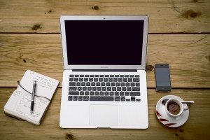 overhead photo of a laptop on a wooden table with notebook, pen, iphone and coffee cup