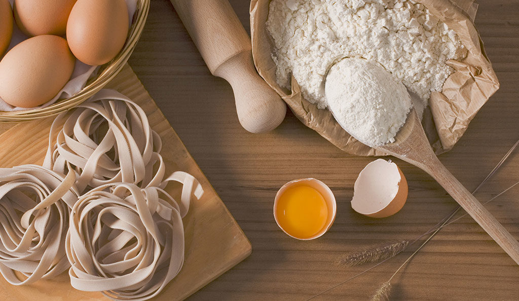 top down view of kitchen counter with utensils and ingredients sitting out: eggs, rolling pin, flour, wooden spoon, pasta
