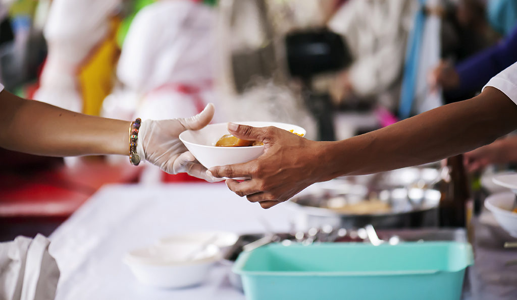 gloved hand of relief worker reaching out bowl of food to a man's hand with other people blurred in background