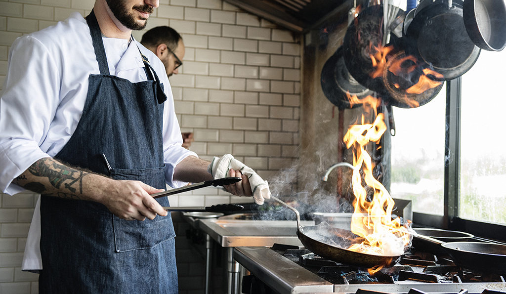 chef with a fiery pan cooking over a kitchen stove in front of a window