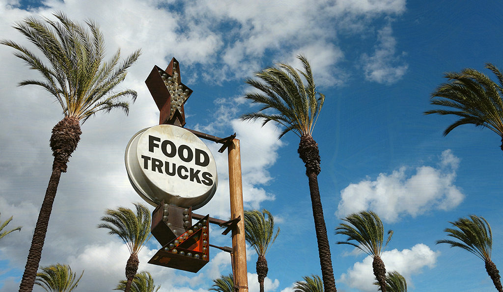 an antique-looking sign surrounded by palm trees that reads "food trucks" with an arrow pointing downward