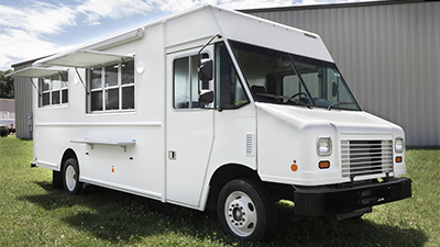 3/4 front angle photo of a white food truck with service windows open parked on grass in front of an industrial building