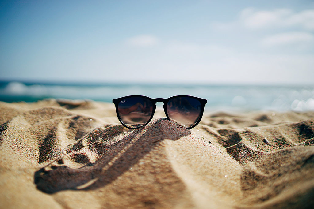 pair of sunglasses sitting on a small mound of sand at the beach with blue water and sky in the background