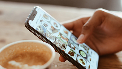 close up photo of a smart phone with the user scrolling through food marketing photos on social media with a cup of coffee in the foreground and wood table top in the background