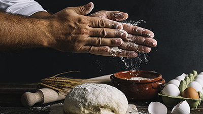 close up of chef hands rubbing flour between them above a ball of dough with various food ingredients in the background