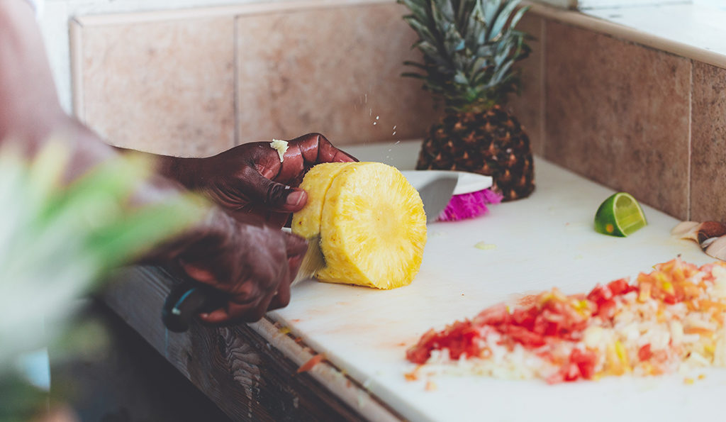 Photograph of black chef's hands cutting up a pineapple with a large knife on a cutting board counter