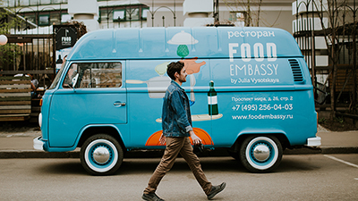 photo of a dark-haired man walking in front of a blue van food truck on a city street