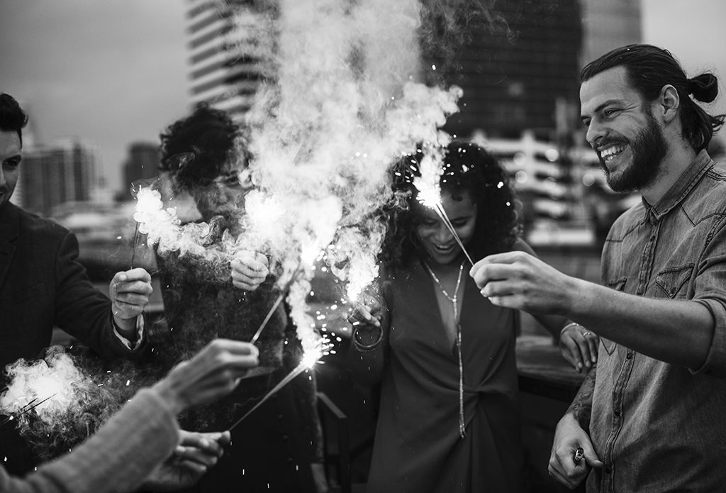 black and white photo of a diverse group of smiling friends lighting sparklers on a city rooftop with buildings in the background