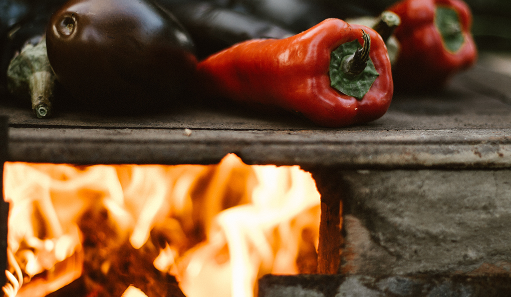 close up photo of red peppers and egg plants being roasted on a grill fire