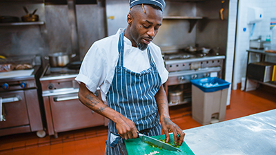 photo of a black man with tattoos on his arms wearing a matching blue-striped apron and chef hat cutting onions in an industrial kitchen setting