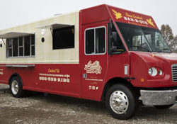 front passenger 3/4 angle photo of a red and cream colored food truck with exterior lights on and serving windows open