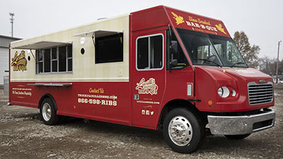 front passenger 3/4 angle photo of a red and cream colored food truck with exterior lights on and serving windows open
