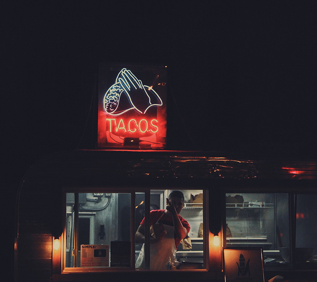 photo at nighttime of a man looking out the window of a food truck with a neon sign on top that says "tacos" with hands holding a taco