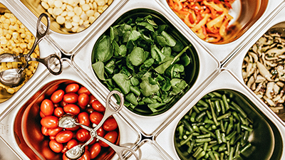 overhead photo of metal sandwich prep food service containers with various vegetables including mushrooms, peppers, tomatoes, corn, green beans and leafy greens