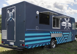 rear three-quarter photo of a light and dark blue food truck with a lion logo on the side parked in a field on a sunny day