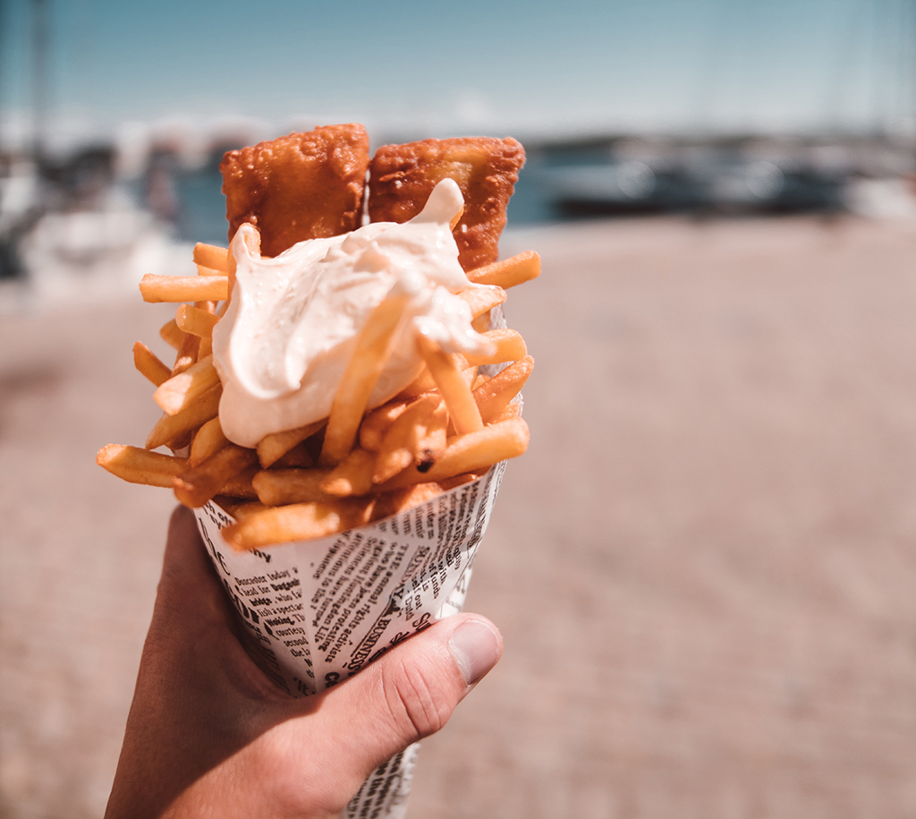 close up of a white person's hand holding a cone of fish and chips with a blurry beach in the background