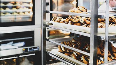 photo of a bakery interior with cinnamon rolls cooling on a baking rack in the foreground and ovens filled with baking goods in the background