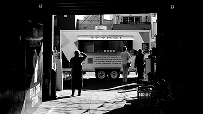 black and white photo looking down a dark alley toward a food truck bathed in light on the street with customers lined up
