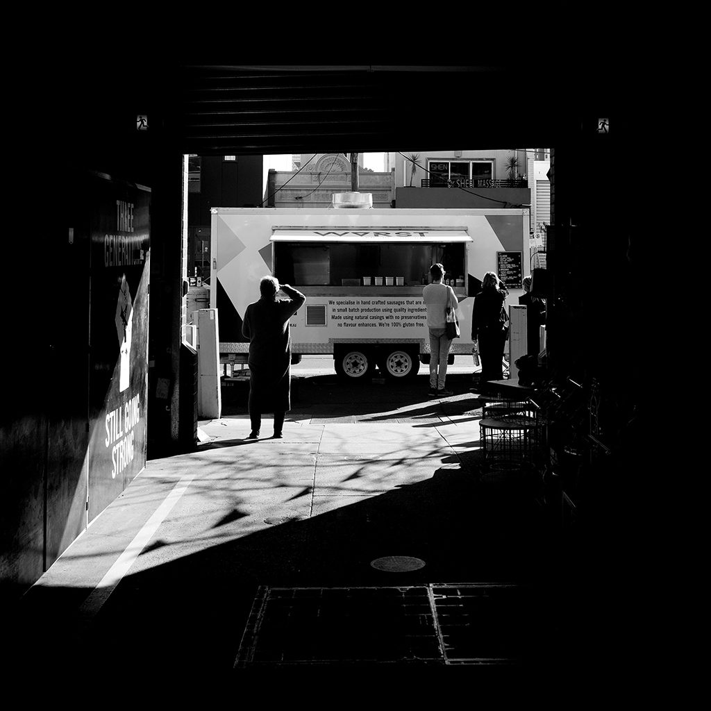 black and white photo looking down a dark alley toward a food truck bathed in light on the street with customers lined up